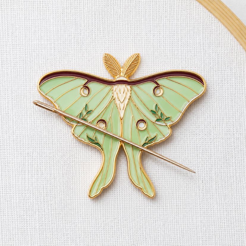 Luna moth enamel needle minder with magnetic back securing a needle on linen, custom sewing notion supplier.