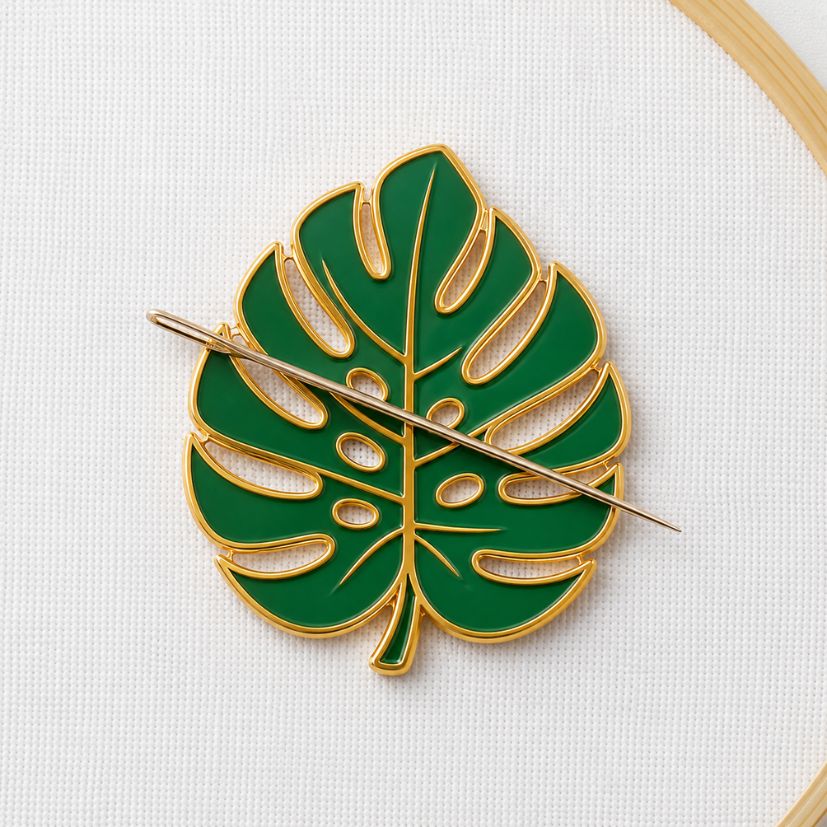 Green monstera leaf enamel needle minder holding a hand sewing needle on hoop fabric, OEM craft merchandise.